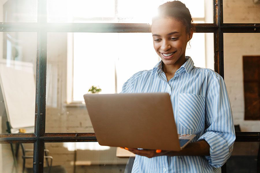 Employee Benefits - Smiling Business Woman Holding a Laptop While Standing in Her Office